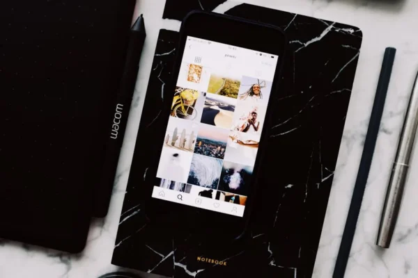 Top view of black notebook with cellphone above with pictures on screen and two pens aside located on marble table